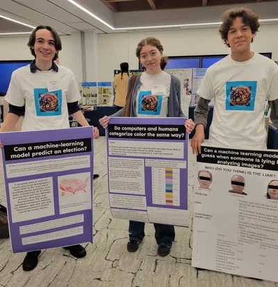 Three students standing behind their research posters at a science fair, presenting projects on whether machine-learning models can predict an election, whether computers and humans categorise colour the same way, and whether a model can detect lying from images