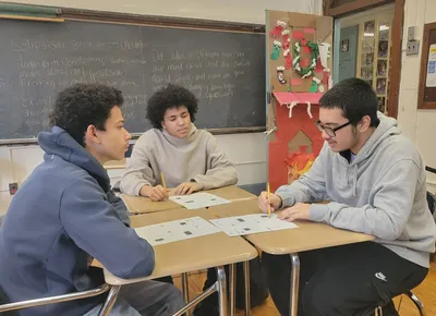 Three students at joined desks discussing a worksheet, with a chalkboard behind them showing notes on the definition of solipsism