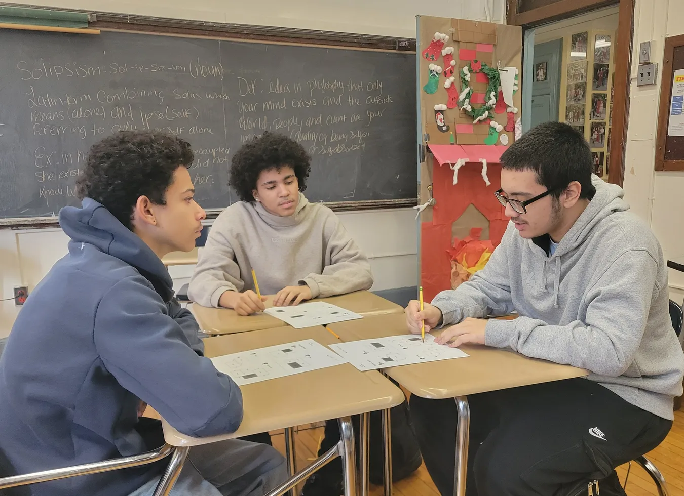 Three students at joined desks discussing a worksheet, with a chalkboard behind them showing notes on the definition of solipsism