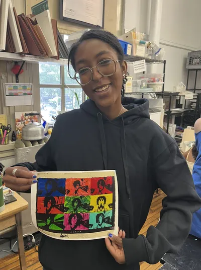 A student in the printmaking studio holding up her nine-panel print, the same portrait repeated in blue, red, yellow, green and pink