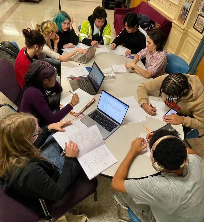 Ten students working around a large round table, some writing in notebooks and others on laptops, in a shared study space