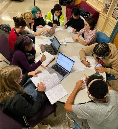 Ten students working around a large round table, some writing in notebooks and others on laptops, in a shared study space