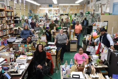 The Urban Academy faculty gathered for a group portrait in their shared staff room, standing and sitting among desks piled with books, papers and potted plants, with student artwork covering the walls behind them