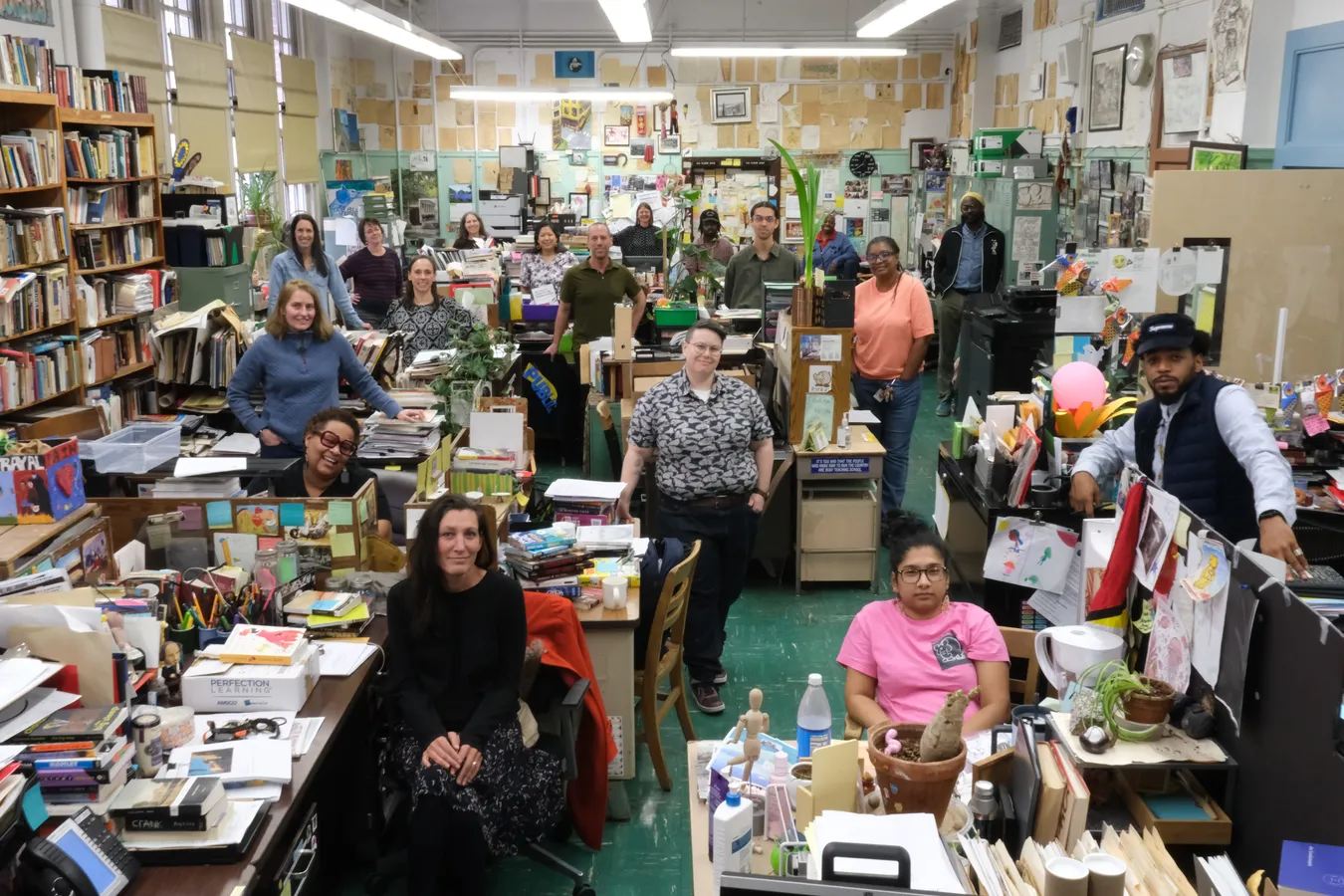 The Urban Academy faculty gathered for a group portrait in their shared staff room, standing and sitting among desks piled with books, papers and potted plants, with student artwork covering the walls behind them