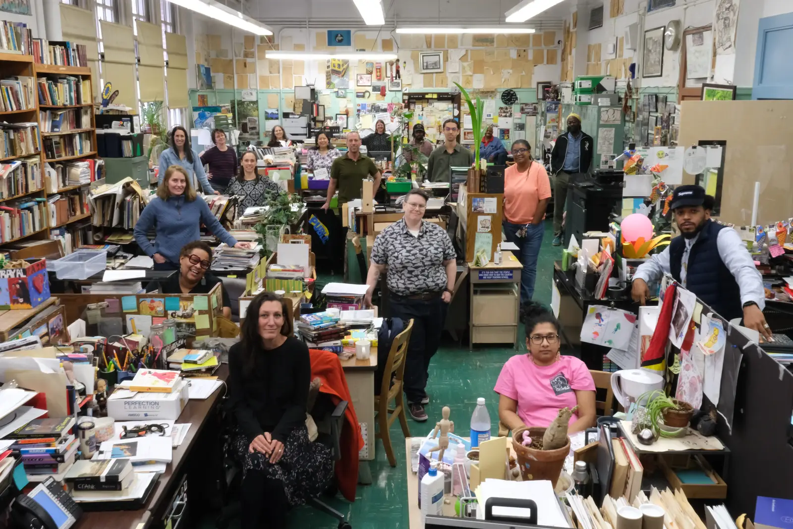 The Urban Academy faculty gathered for a group portrait in their shared staff room, standing and sitting among desks piled with books, papers and potted plants, with student artwork covering the walls behind them