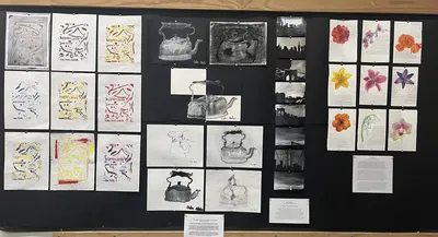 A wall of student work mounted on black paper: relief prints in Cyrillic lettering, charcoal and pencil studies of a kettle, black-and-white photographs of New York bridges, and watercolour flower studies annotated by hand