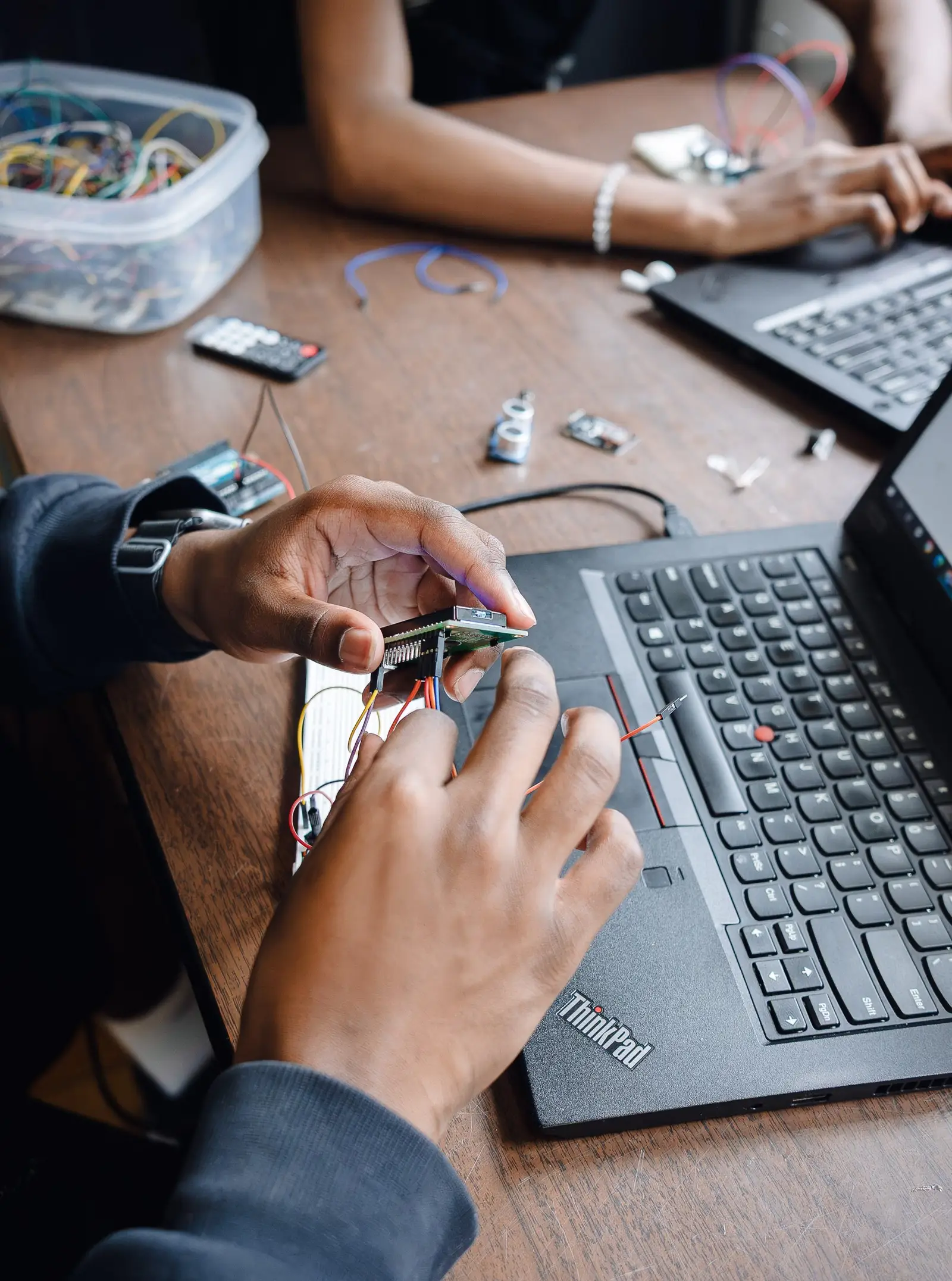 Close-up of a student's hands connecting jumper wires to a small circuit board resting on a laptop keyboard, while another student works at a second laptop across the table