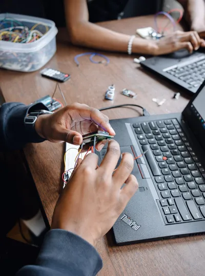 Close-up of a student's hands connecting jumper wires to a small circuit board resting on a laptop keyboard, while another student works at a second laptop across the table