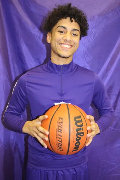 A student smiling and holding a basketball in a purple JREC warm-up jacket, photographed against a purple backdrop
