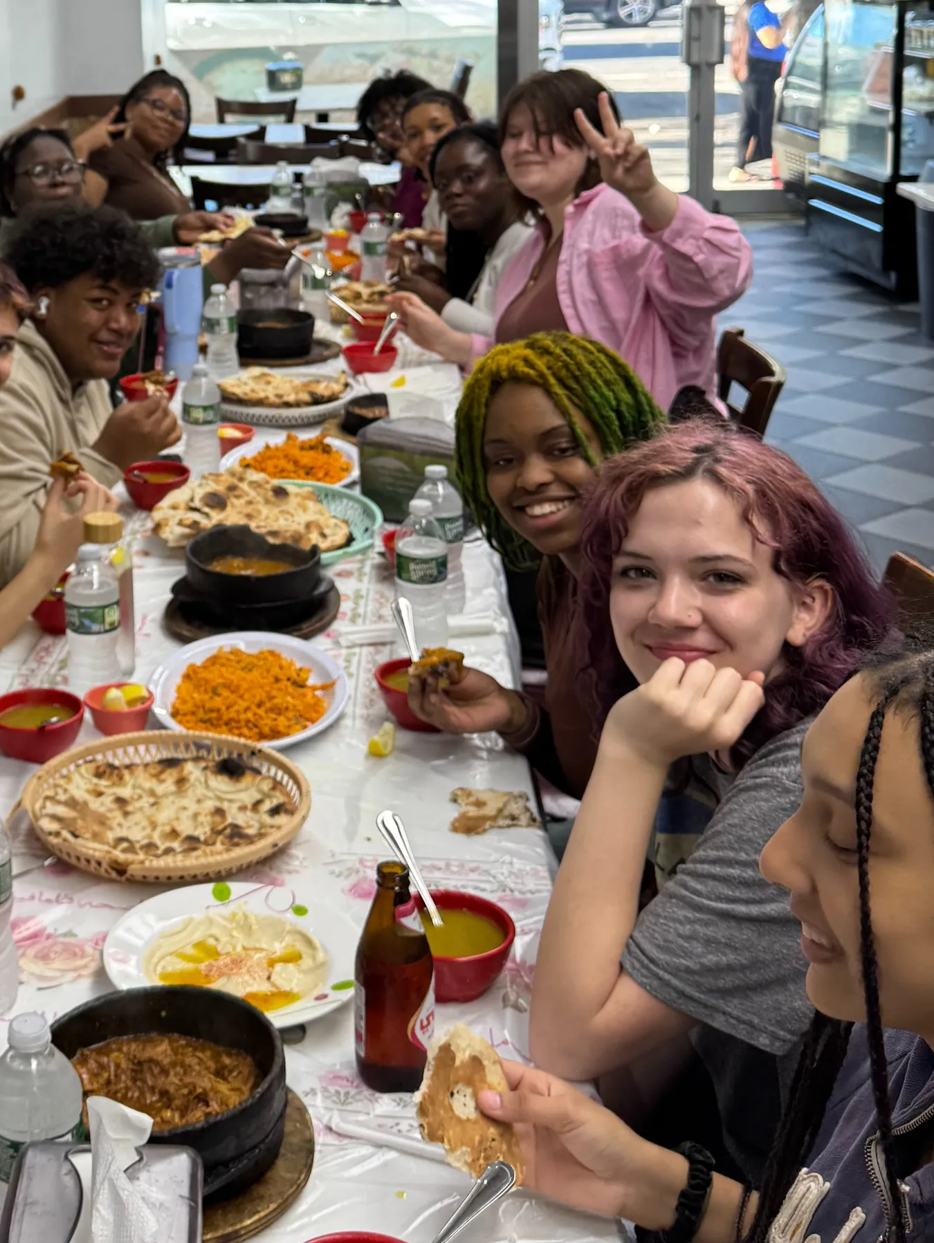 Students gathered around a long restaurant table piled with naan, orange rice and stews in small cast iron pots, several smiling for the camera and one flashing a peace sign, during a food-tasting trip