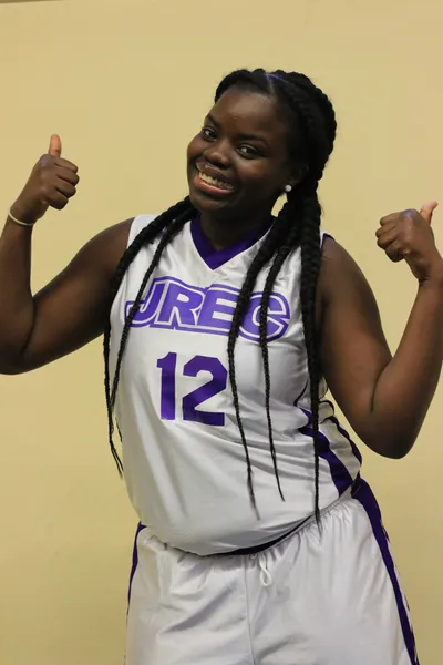 A student athlete smiling and pointing to the number 12 on the back of her white JREC jersey, her braided hair swept over one shoulder