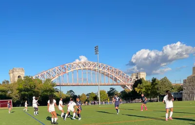 JREC girls soccer players in white and navy jerseys converging on the ball during a match on a turf field, a large steel arch bridge rising behind the trees along the water