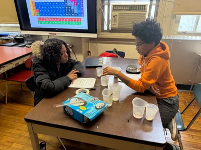 Two students setting up plastic cups for an experiment on a lab bench, with a periodic table displayed on the screen behind them