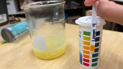 A hand holding a pH test strip labelled "lemon" against the colour chart on the tube, beside a beaker of cloudy yellow liquid on a lab bench