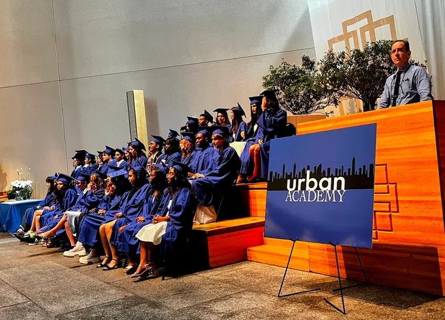 Graduates in blue caps and gowns seated on tiered steps beside a lectern and an Urban Academy sign, with a staff member speaking at the podium