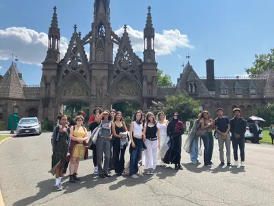 A class of students posing together on a trip, standing in front of the Gothic revival stone arch at the entrance to Green-Wood Cemetery
