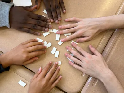 Six students' hands reaching in from around a table, sorting small blank paper tiles into groups during a sorting activity