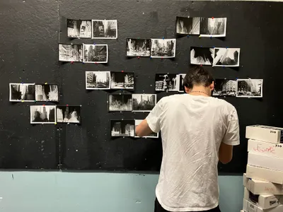 A student pinning black-and-white photographs of New York City buildings onto a large dark display board, with boxes of photographic paper stacked beside them
