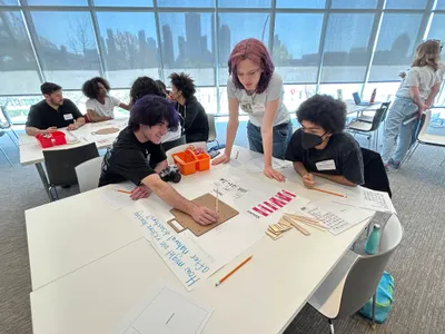 Students working around a table on a design-build task with craft sticks and a pegboard, a teacher leaning in to help, beside a handwritten prompt asking how many people a design affects, in a bright room overlooking a city skyline