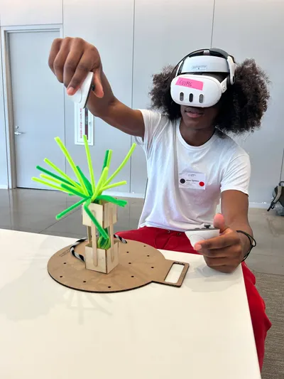 A student wearing a virtual reality headset and holding a controller, reaching out towards a wooden model topped with bright green rods