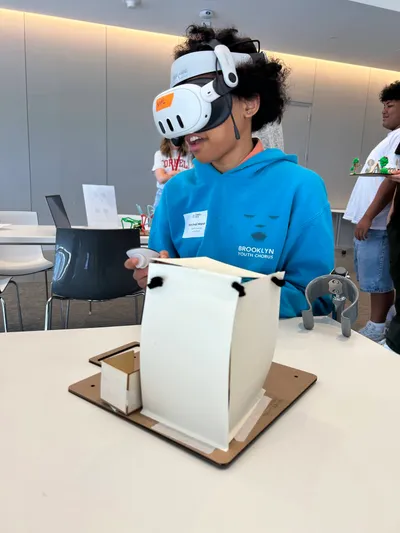 A student in a VR headset reaching towards a white card model mounted on a wooden base during a design workshop