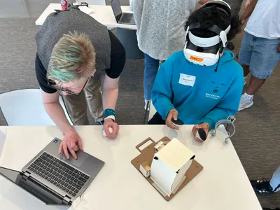 A workshop leader working at a laptop while a student in a VR headset handles the physical model beside them, seen from above
