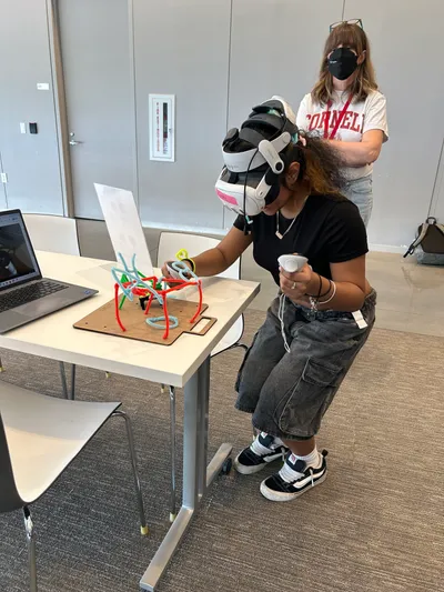 A student wearing a VR headset reaching towards a model built from coloured pipe cleaners, while an instructor watches from behind