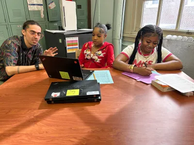 A teacher talking with two students across a table, a laptop open between them and worksheets and books spread out in front of the lockers