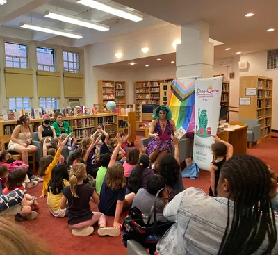 Urban Academy students helping run a Drag Queen Story Hour in a library, with young children sitting on the floor raising their hands to answer the reader's question