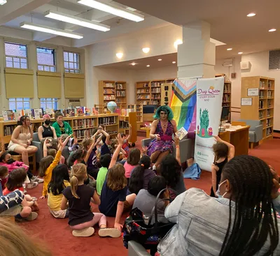 Urban Academy students helping run a Drag Queen Story Hour in a library, with young children sitting on the floor raising their hands to answer the reader's question