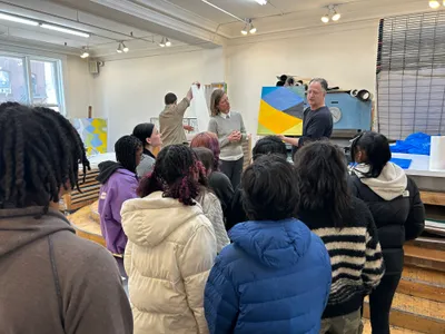 A class of students gathered in an artist's studio listening to two artists talk about a large yellow and blue abstract canvas