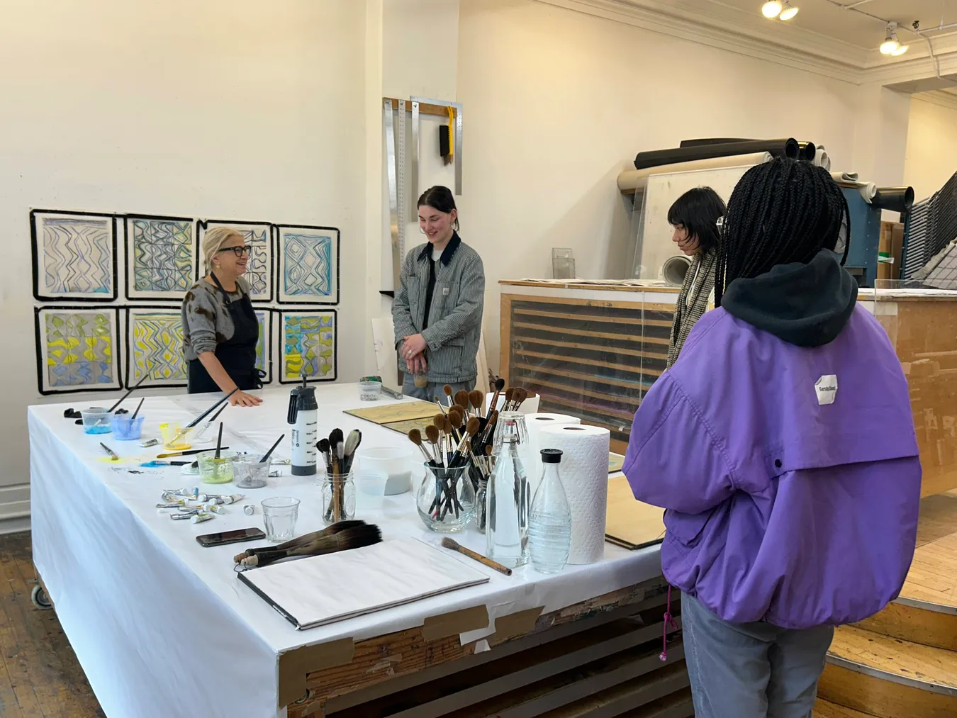 Students visiting a printmaking studio, listening to the printers beside a table of brushes and inks with a grid of finished prints on the wall behind