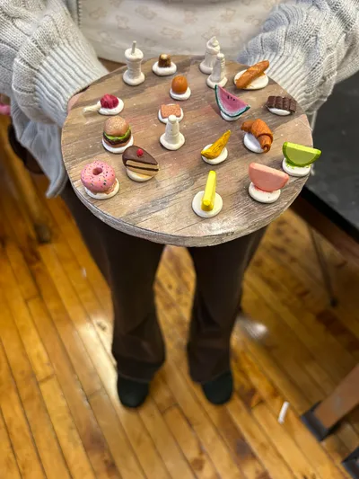 A student holding out a round wooden board arranged with handmade clay miniatures of food, including a burger, a slice of cake, a doughnut, a croissant and a wedge of watermelon