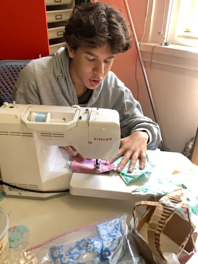 A student feeding patterned fabric through a Singer sewing machine, surrounded by fabric scraps and materials