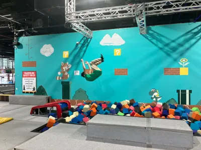 A student caught mid-somersault in the air above a foam pit at an indoor trampoline park, against a brightly painted wall