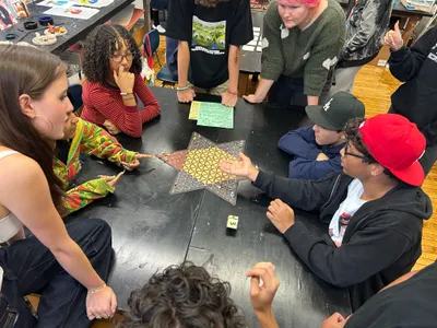 Students gathered around a table in an art room playing a star-shaped board game together, one gesturing as he explains a move