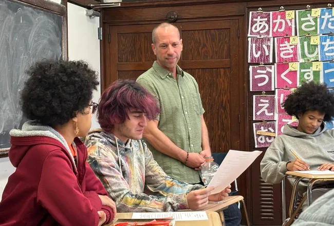 A teacher standing beside students at their desks while one reads aloud from a printed handout, in a classroom with Japanese hiragana charts on the wall