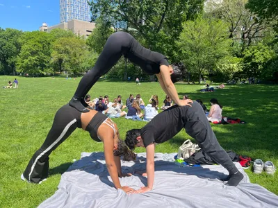 Two students holding a paired acro-yoga pose on a blanket in Central Park, with classmates sitting on the grass behind them