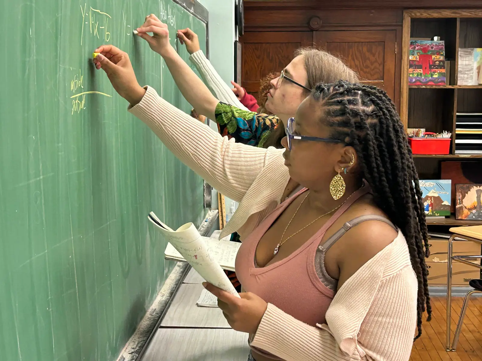 Two students working through an equation on a green chalkboard, one writing while the other holds an open notebook and checks her working