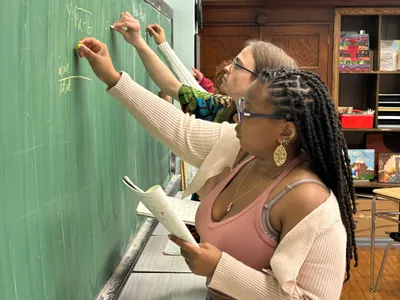 Two students working through an equation on a green chalkboard, one writing while the other holds an open notebook and checks her working