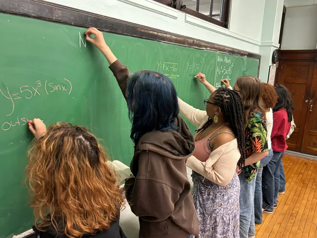 A row of students standing at a green chalkboard, each working through a different calculus problem