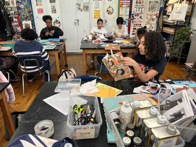 Students working on collages at tables in the art room, one holding up a finished piece, with trays of scissors, glue and cut paper in the foreground