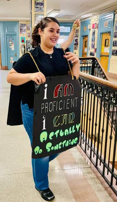 A student ringing a small brass bell overhead while wearing a hand-lettered sandwich board reading "I am proficient, I can actually do something", on a school landing lined with framed photographs