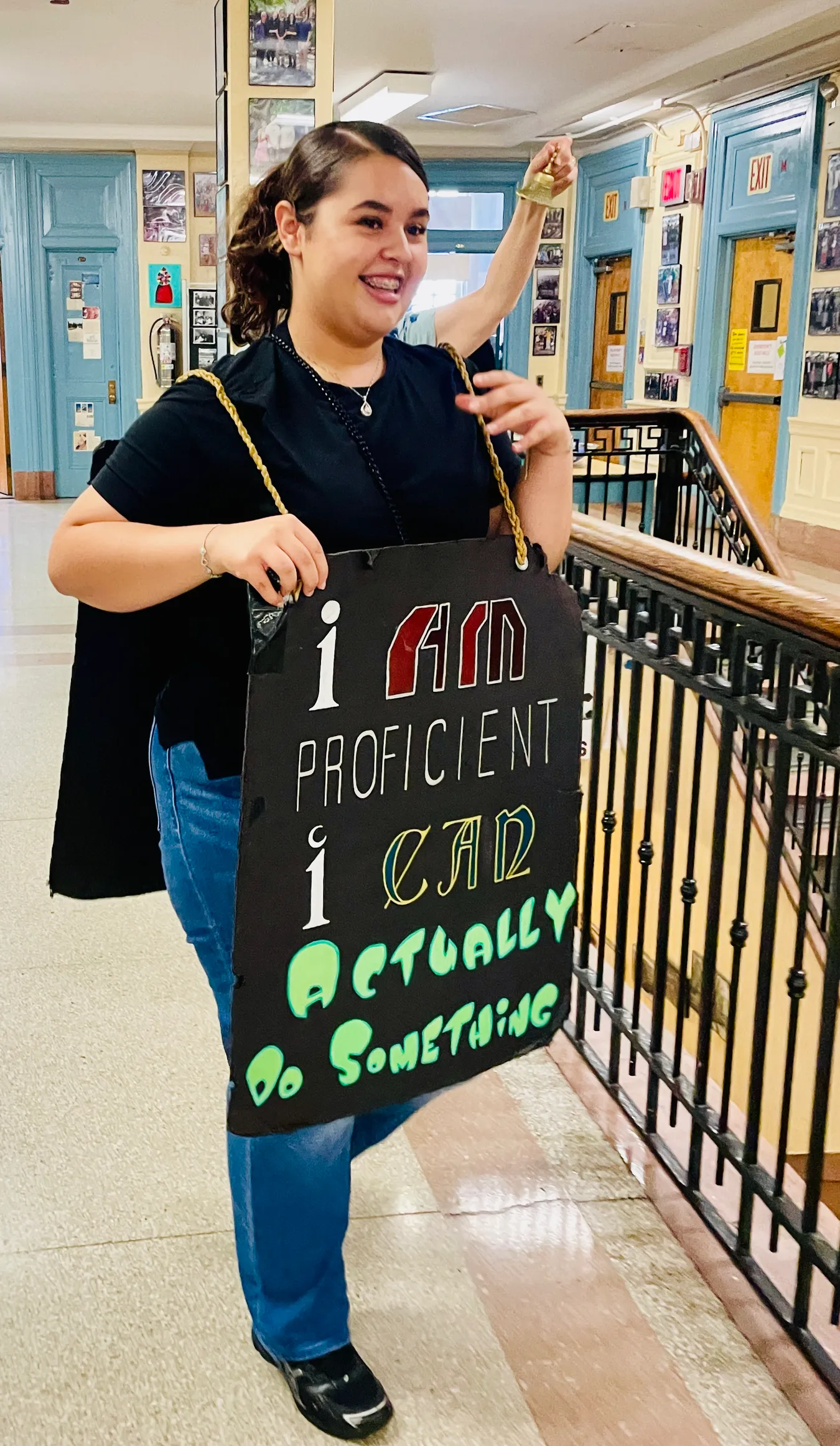 A student ringing a small brass bell overhead while wearing a hand-lettered sandwich board reading "I am proficient, I can actually do something", on a school landing lined with framed photographs