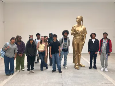 A class of students posing beside a life-size gold figurative sculpture in a bright white gallery on a museum trip