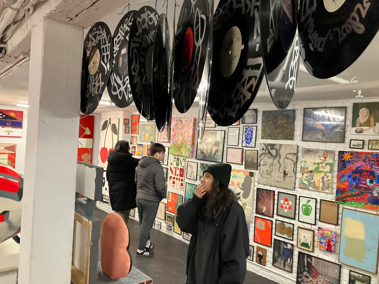 Students in an art space looking at a salon-style wall hung edge to edge with paintings, prints and framed work, with graffiti-tagged vinyl records suspended from the ceiling above them