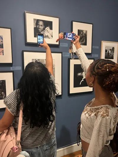 Two students photographing framed black-and-white portraits on a blue-grey gallery wall with their phones