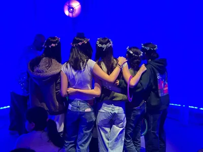 A group of students in flower crowns standing arm in arm with their backs to the camera, lit by deep blue light at a school event