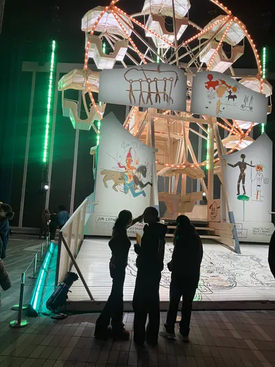 Students silhouetted in front of a large illuminated art installation of a wooden ferris wheel hung with painted panels