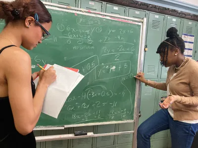 Two students working through systems of linear equations on a green chalkboard, one writing on the board while the other checks her notes